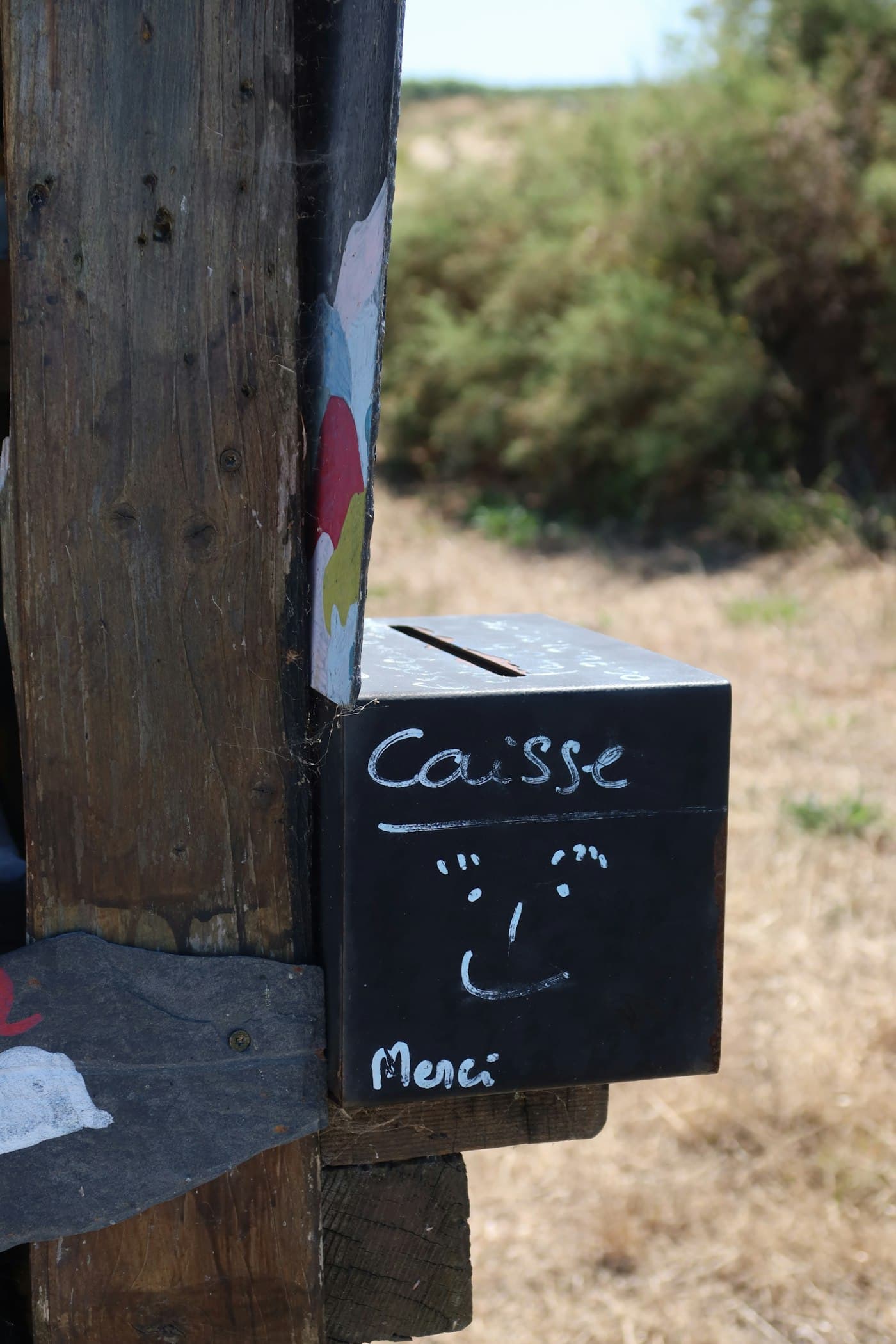 A black metal cash box on a wooden post, hand-lettered “Caisse” and “Merci” with a smiley face.