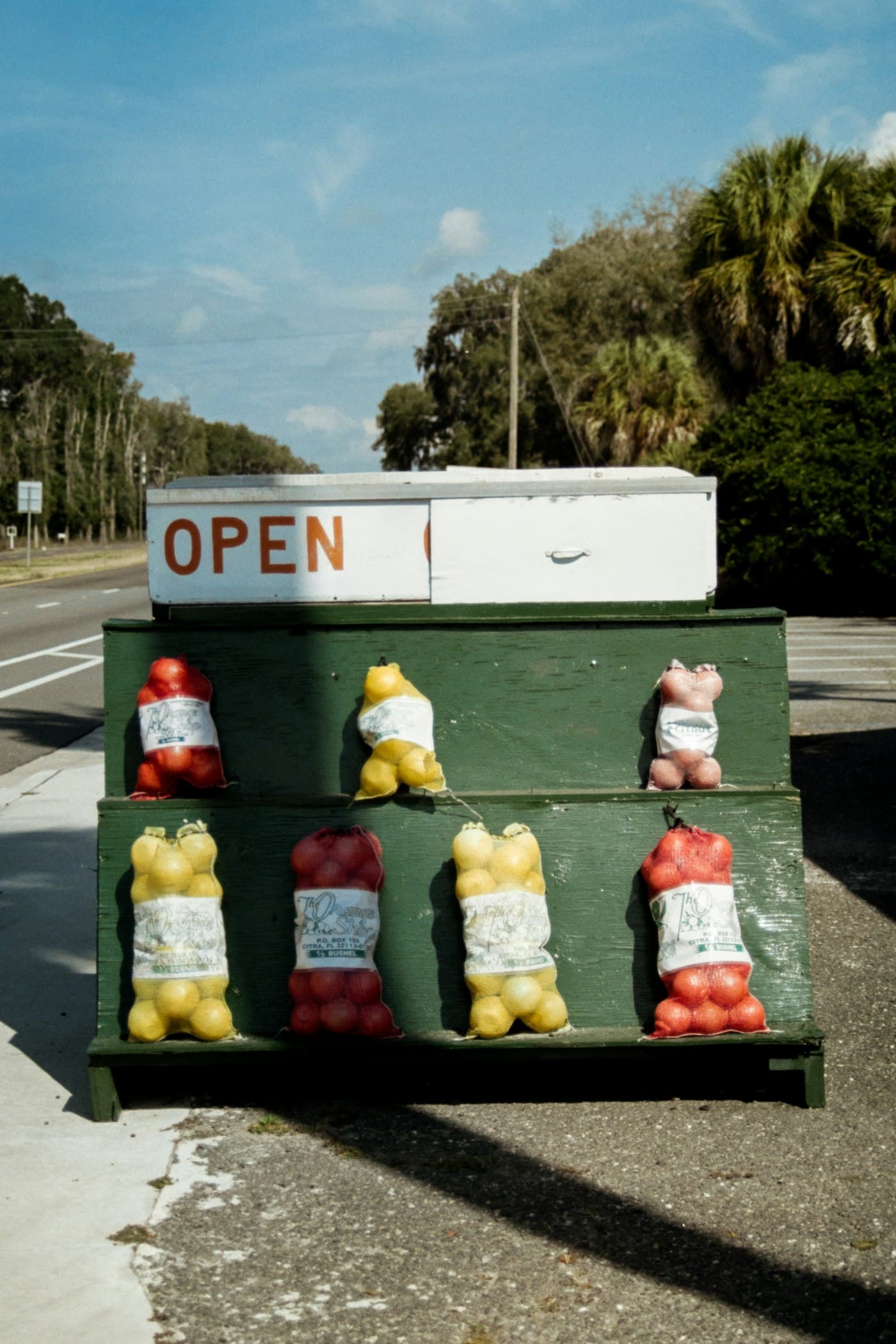 A roadside produce stand with an OPEN sign and mesh bags of citrus on green wooden shelves, nobody attending it.